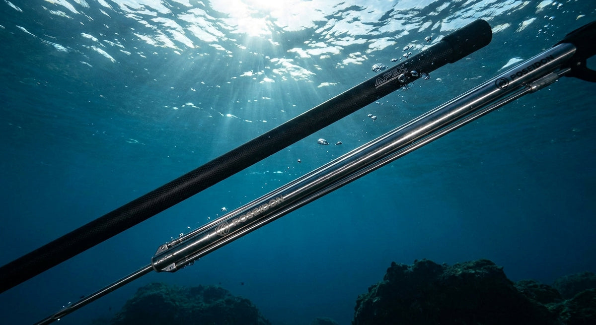 Carbon fiber pole spear and stainless steel speargun shaft suspended underwater with sunlight refracting through clear blue ocean water above.